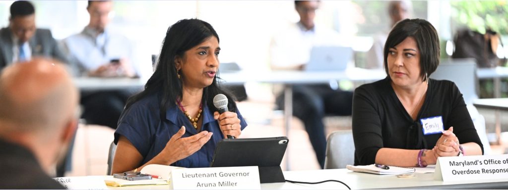 A picture of Maryland Lt. Governor Aruna Miller and Maryland Special Secretary of Overdose Response Emily Keller sitting behind a table. Lt. Governor Miller is speaking into a microphone.
